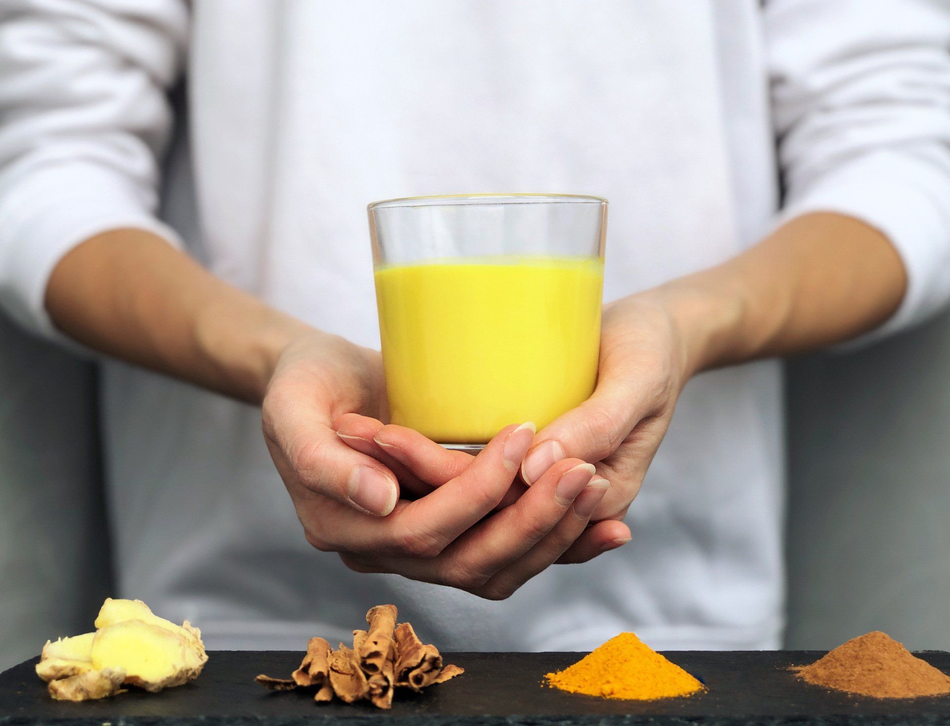 Hands holding a glass of yellow juice above spices and ginger on a black surface