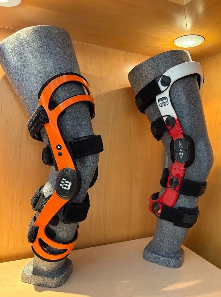 Two knee braces on gray mannequin legs, one orange and black, one red and black, displayed on a shelf.