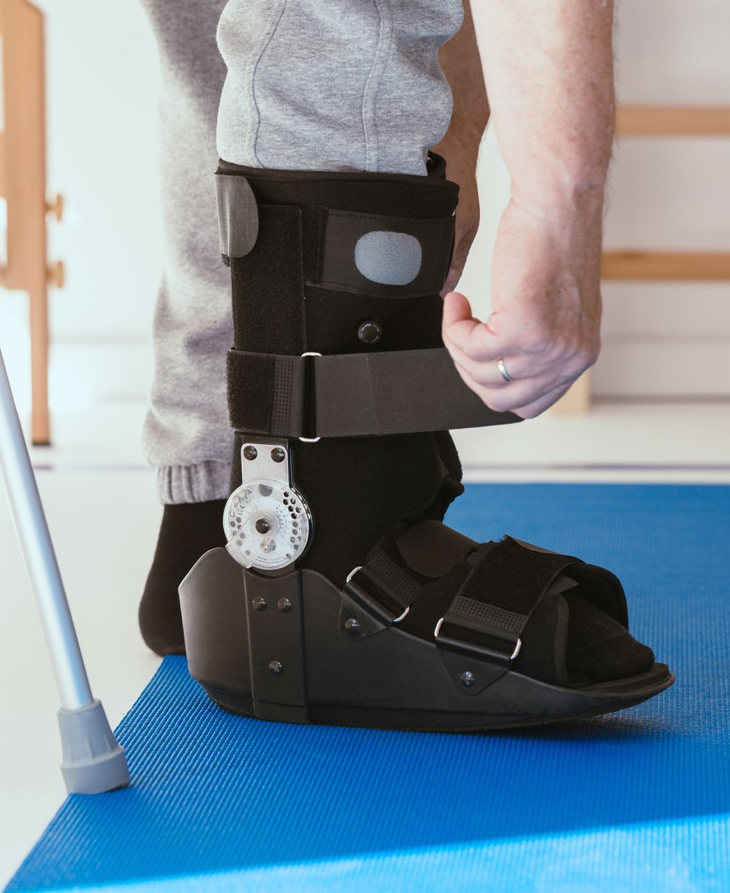 Person wearing a black ankle-foot brace and white boot in a clinical setting, standing on a blue floor mat