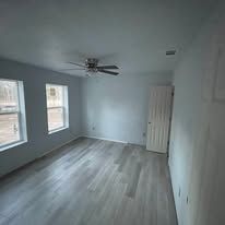 Empty gray bedroom with wood-look floor, two windows, ceiling fan, and white door.