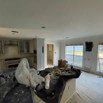 Bright living room with plastic-covered furniture, open kitchen, and large windows looking outside