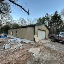 Tan metal building with garage door beside a pickup truck, with debris piled in the muddy foreground