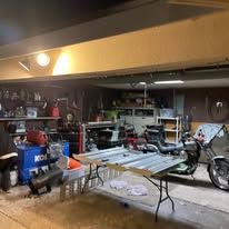 Garage/workshop with a blue machine, tools, tables, and a motorcycle under bright overhead lights.