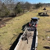 Flatbed truck carrying debris along a dirt path beside a fenced, grassy field with bare trees