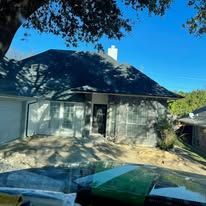 Front view of a gray house with a dark roof, tree shadows, and cars visible at the bottom edge.