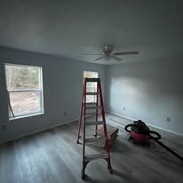 Empty room under renovation with a red ladder, ceiling fan, and a floor tool near the wall.