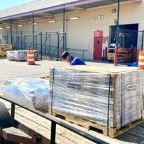 Wrapped pallets on a cart outside a warehouse loading dock, with a worker crouching nearby.