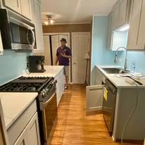 Narrow kitchen with white cabinets and wood floor, with a person standing in the doorway.
