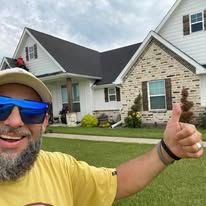 Smiling man in yellow shirt gives a thumbs-up in front of a white house with brick siding.