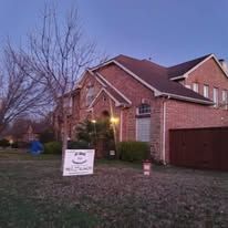 Brick suburban house at dusk with a lit porch, leafless tree, and a sign in the front yard