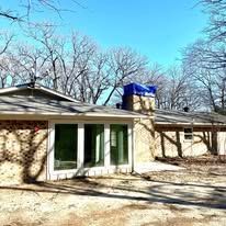 Single-story beige house with white-framed glass doors and a blue roof vent under leafless trees.