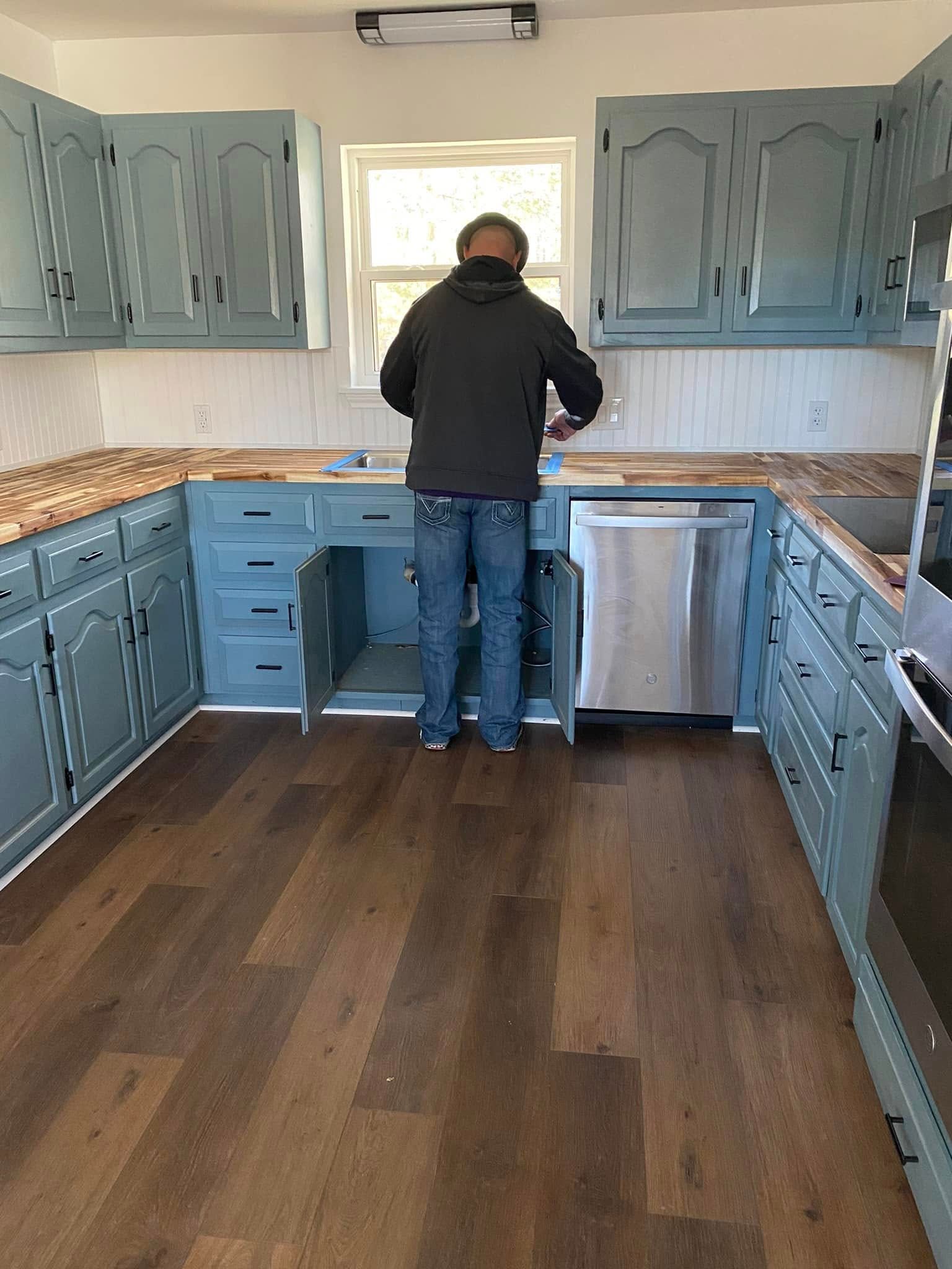 Person standing in a blue kitchen, facing the sink with cabinet doors open and hardwood floor visible.