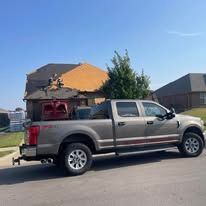 Silver pickup truck parked on street beside damaged house with roof torn off, debris visible.