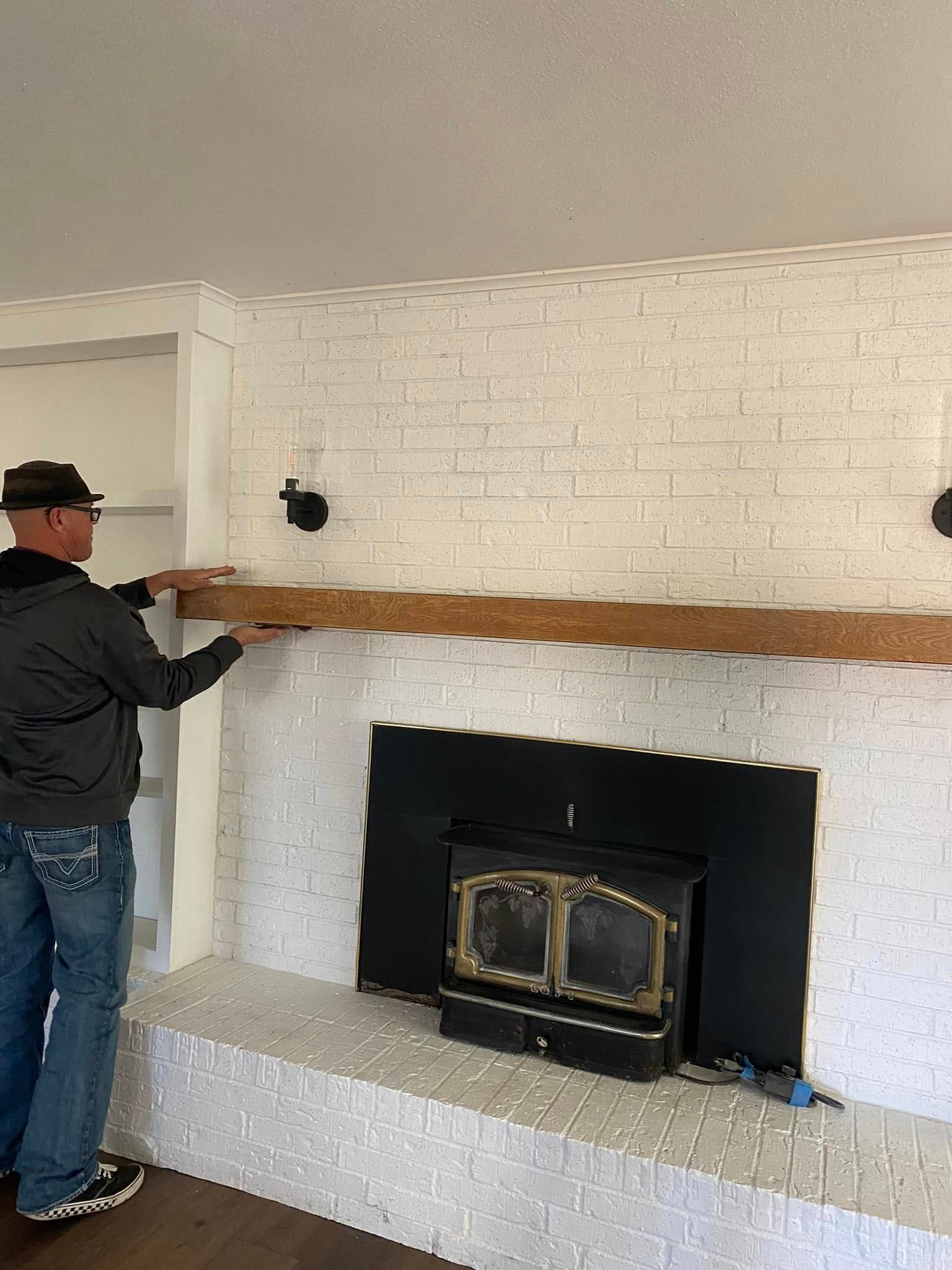 Person installing a rustic wood mantel above a black fireplace in a white brick living room