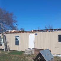 Single-story beige mobile home with white door, ladder, and tarp-covered roof under a clear blue sky
