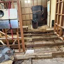 Damaged wooden floor in a room under renovation, with exposed joists and a fan near the wall.