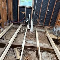 Exposed floor joists and studs in a room under renovation, with wiring and plumbing visible.