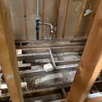Open bathroom floor with exposed wooden joists, plumbing pipes, and rough construction debris