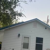 White house side with blue trim, a window, vent, and trees along the roofline.