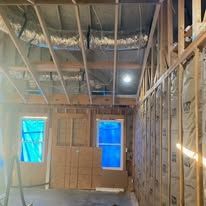 Unfinished room with exposed framing, insulation, and two covered windows, lit by ceiling lights.