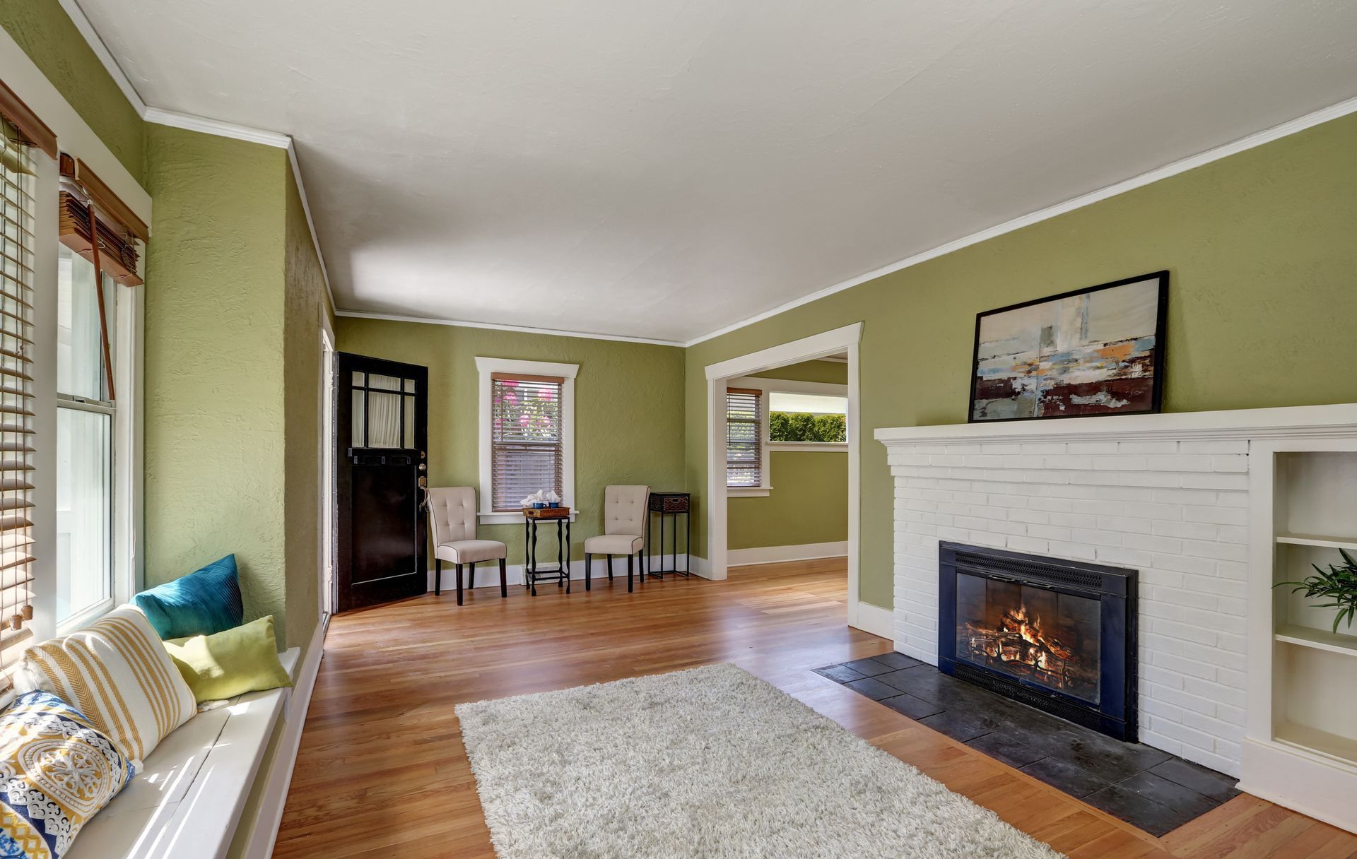 Bright living room with green walls, white fireplace, hardwood floors, and large window seating.