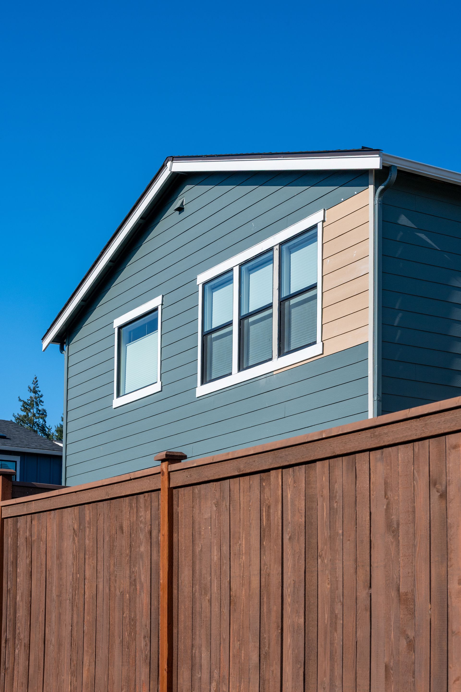 Blue-gray house with white-trimmed windows behind a tall wooden fence under a clear blue sky