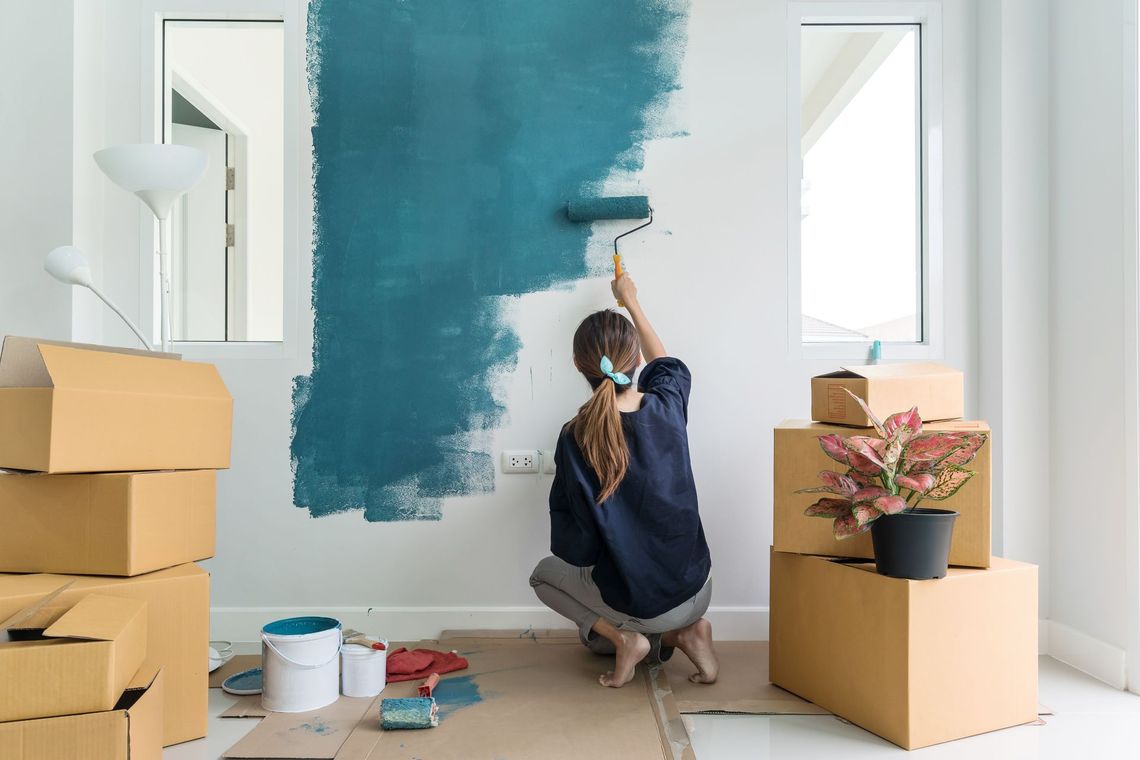 Person painting a teal accent wall in a bright room with boxes and supplies nearby