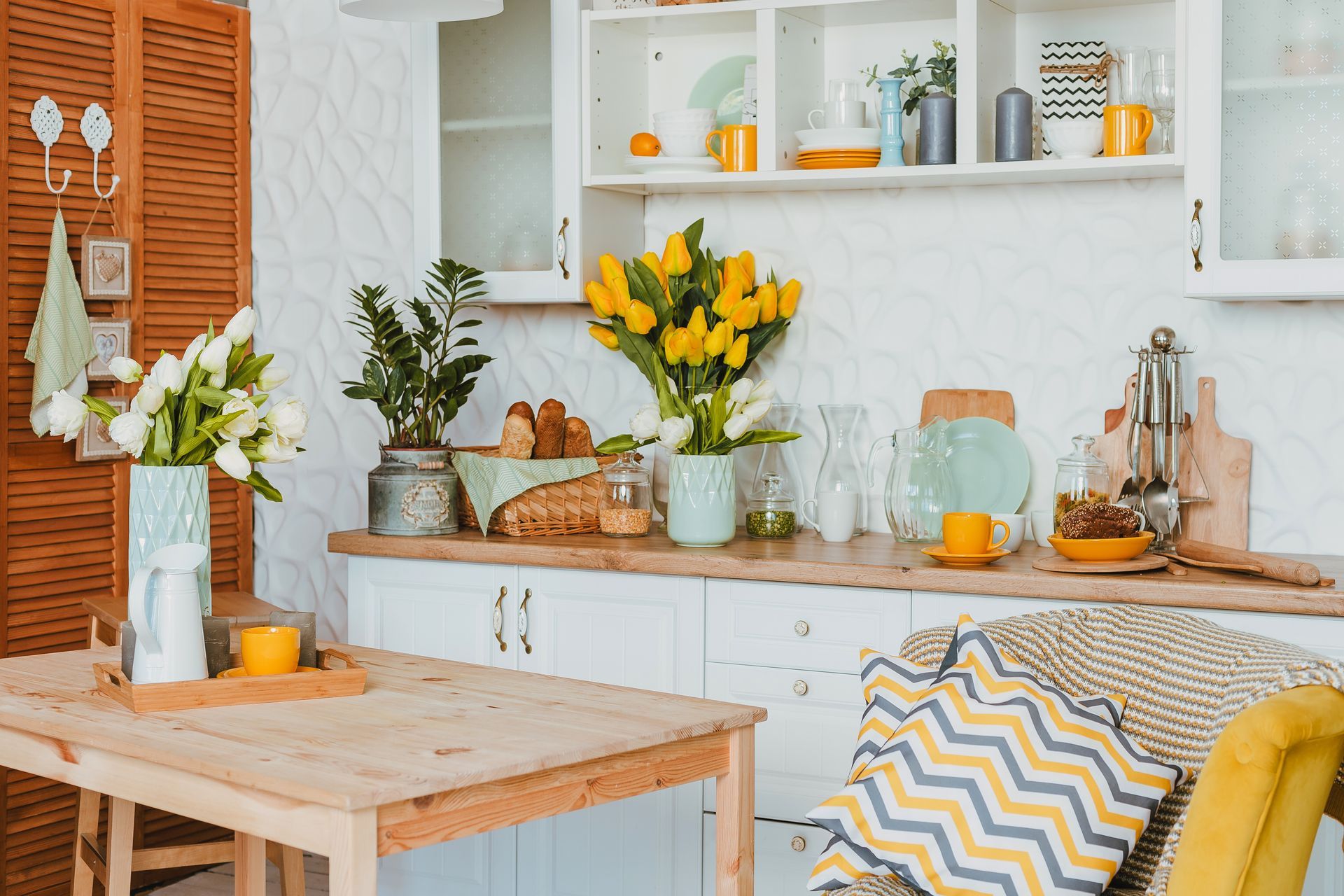Bright kitchen with white shelves, yellow flowers, wooden table, and woven chair with patterned cushion