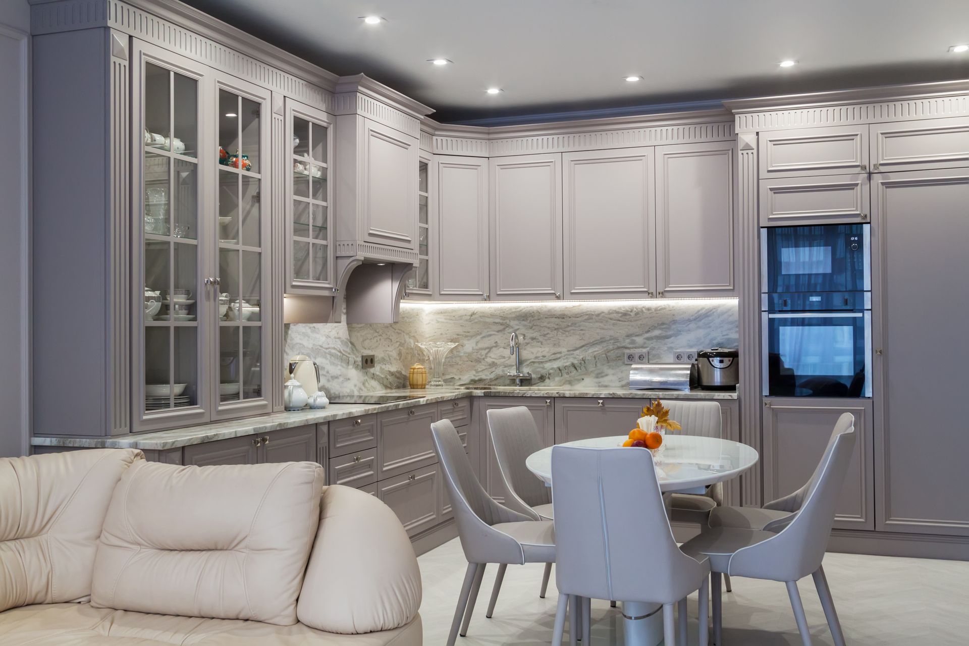Bright modern kitchen with gray cabinets, glass-front storage, a round dining table, and a beige sofa.