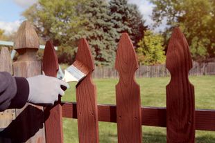 Gloved hands painting a red wooden fence in a garden with trees and grass in the background