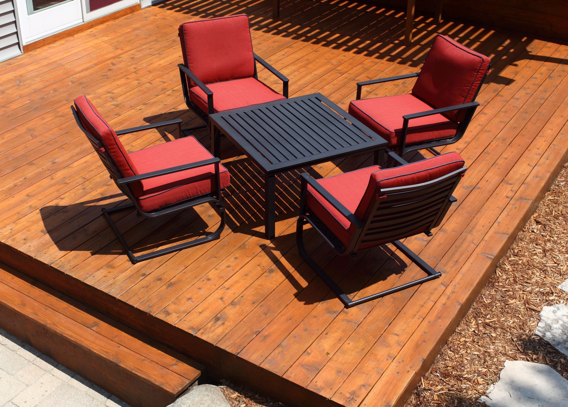 Four red chairs around a black table on a wooden deck in sunlight