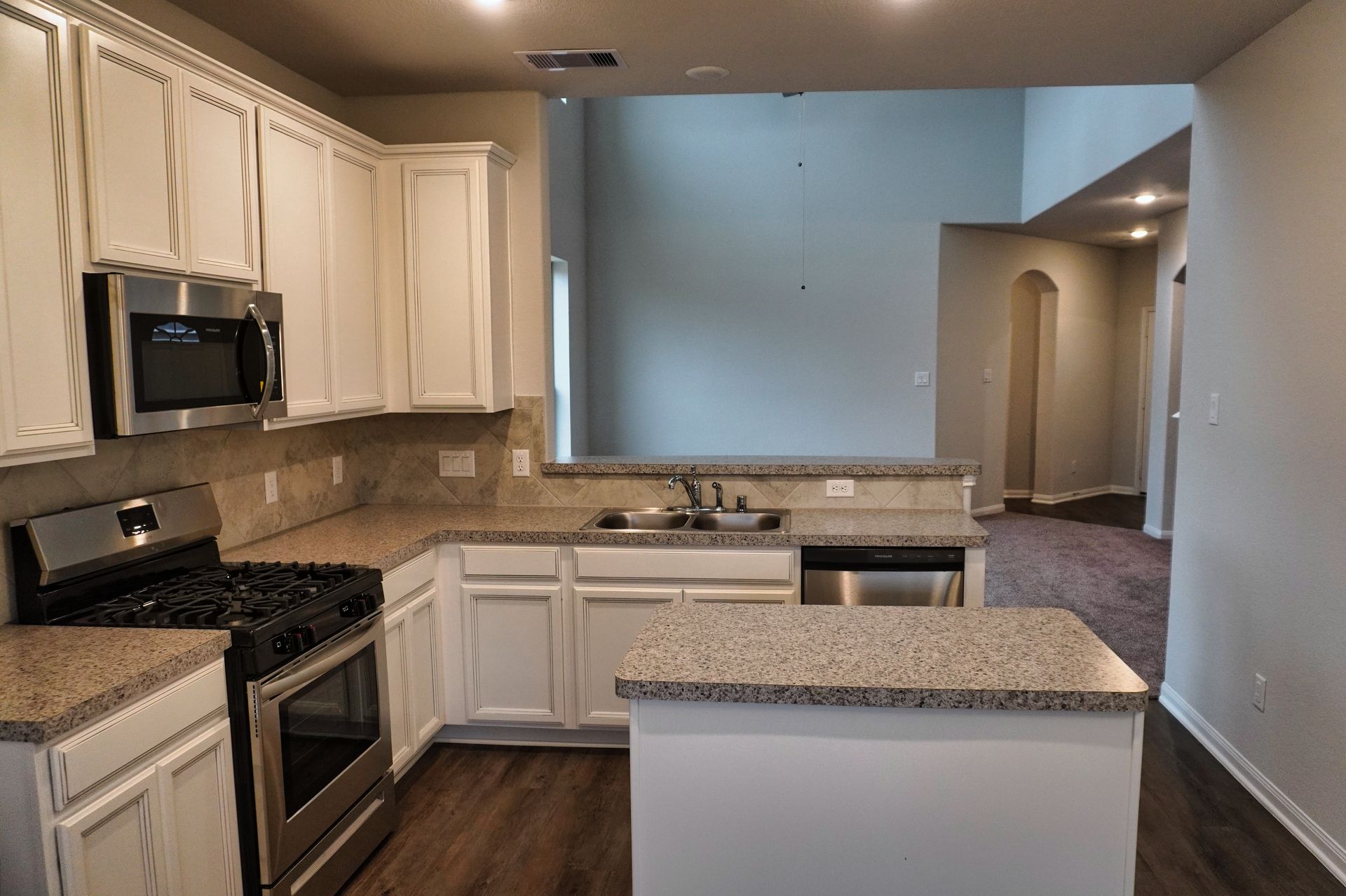 Empty kitchen with white cabinets, granite counters, and a peninsula opening to a carpeted hallway.