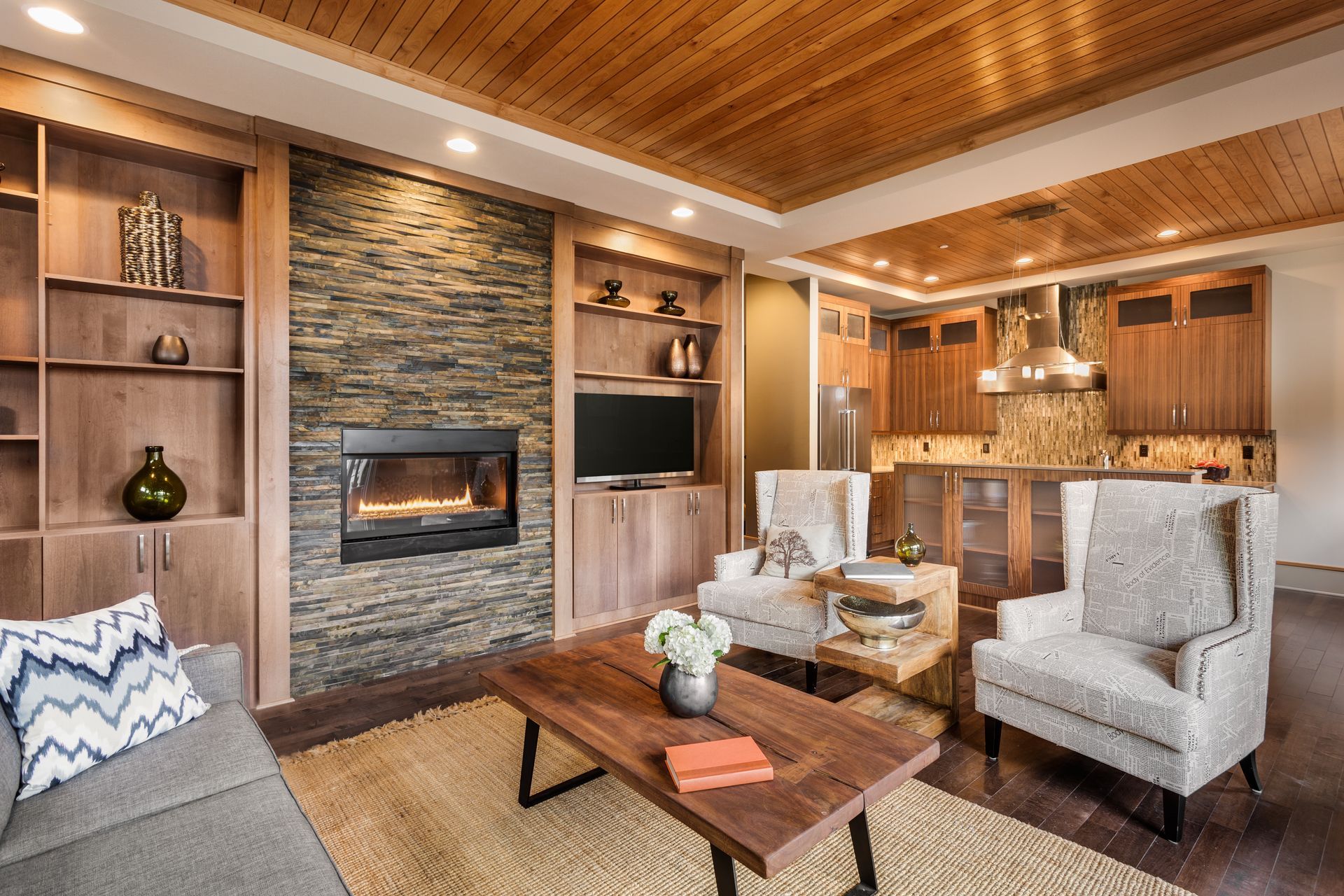 Cozy living room with stone fireplace, wood ceiling, beige chairs, and open kitchen area