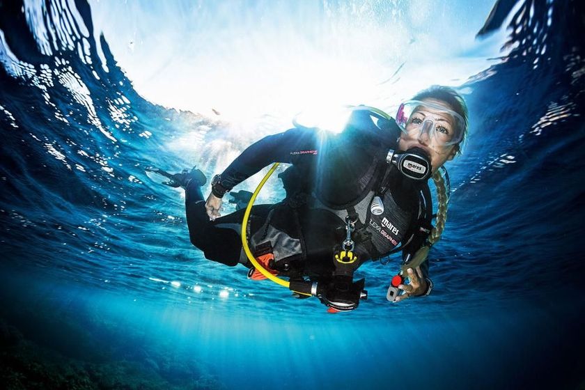 Snorkeler in black wetsuit surfacing in water, wearing mask and raising an OK hand signal.