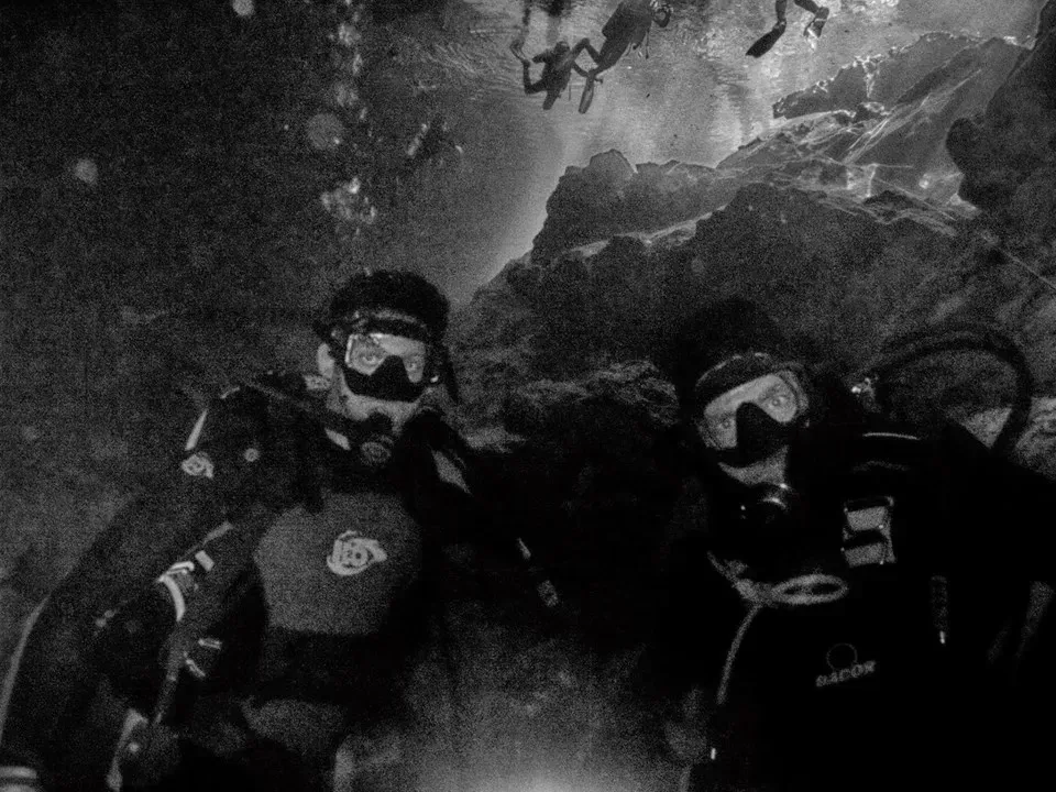 Two scuba divers in masks and wetsuits underwater near a rocky wall