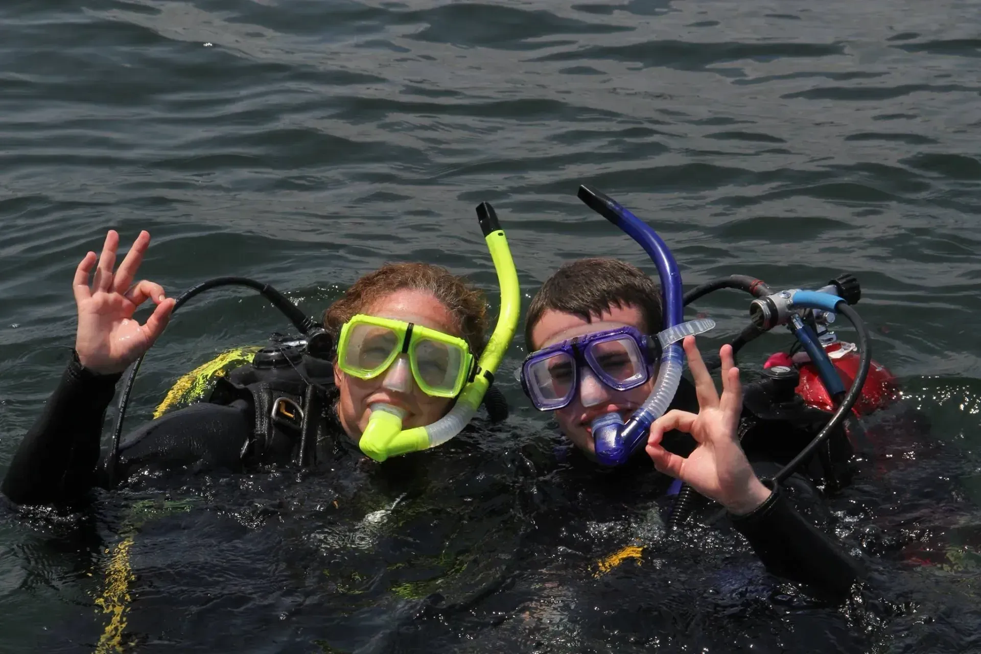 Two snorkelers in black wetsuits float in the water, wearing green and blue masks and raising OK signs.