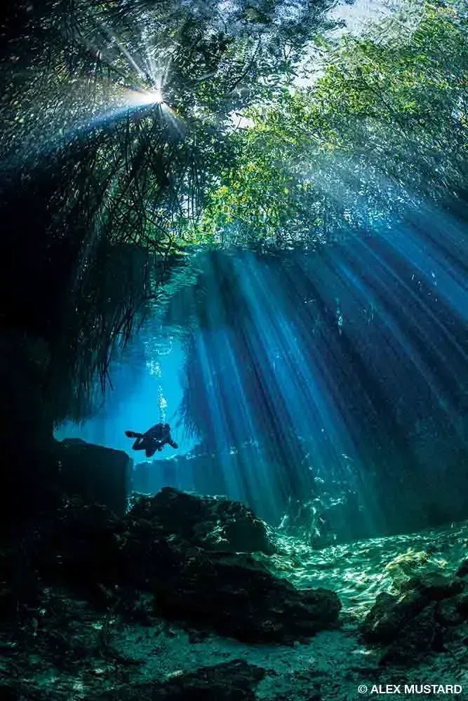 Sunbeams streaming into a turquoise underwater cave with a lone diver silhouetted below