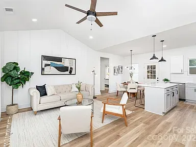 Bright open living room and kitchen with white walls, wood accents, sofa, chairs, and ceiling fan.