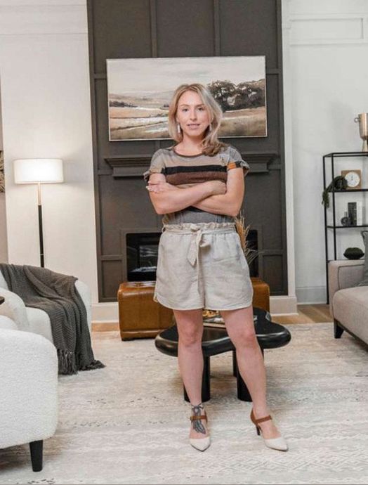 Woman standing in a living room, arms crossed, wearing a striped top and beige shorts.