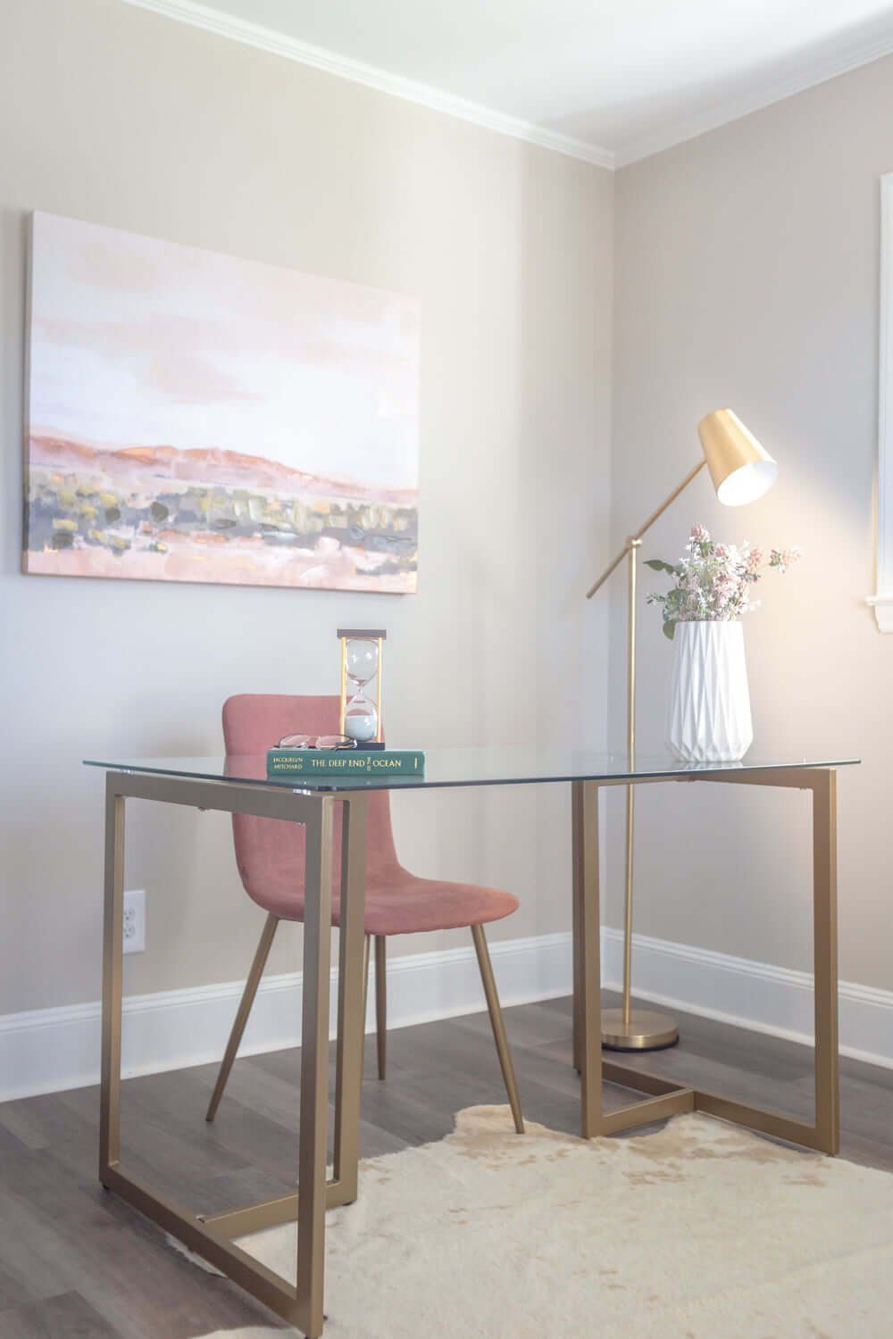 Small home office with glass desk, pink chair, lamp, flowers, and wall art in a corner room.