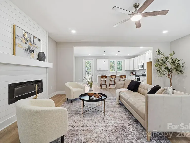 Bright modern living room with white fireplace, beige sofa, chairs, coffee table, and ceiling fan