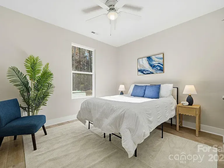 Bright bedroom with a white bed, blue chair, bedside tables, and a large plant by the window