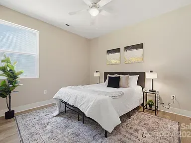 Bright bedroom with a made bed, nightstand lamp, window, and potted plant on a gray rug.