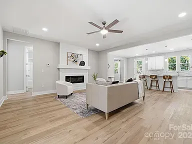 Bright open living room with white fireplace, gray sofa, wood floors, and kitchen bar stools nearby