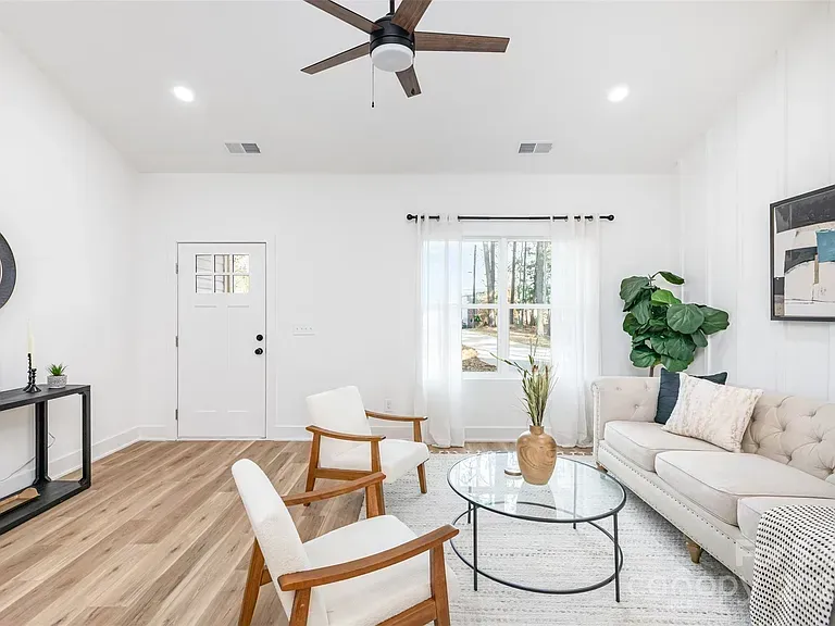 Bright living room with white sofa, glass coffee table, wooden chairs, ceiling fan, and natural light from sliding doors