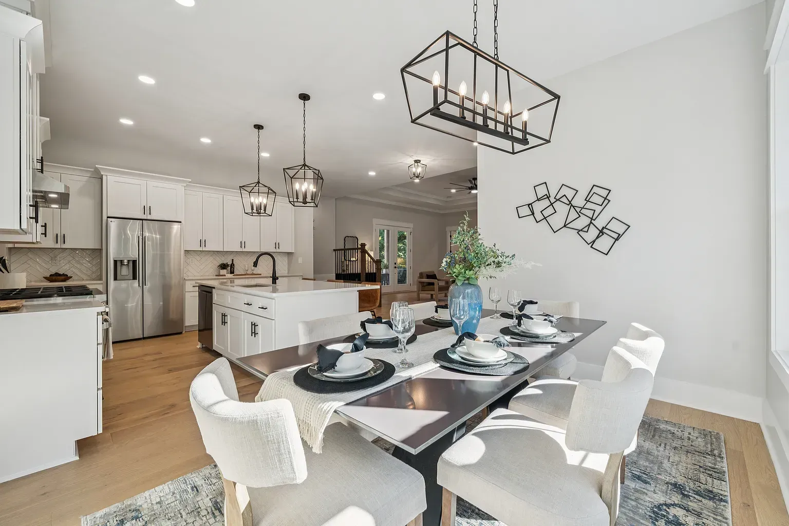 Bright open dining room and kitchen with white chairs, a set table, island, and modern pendant lights.