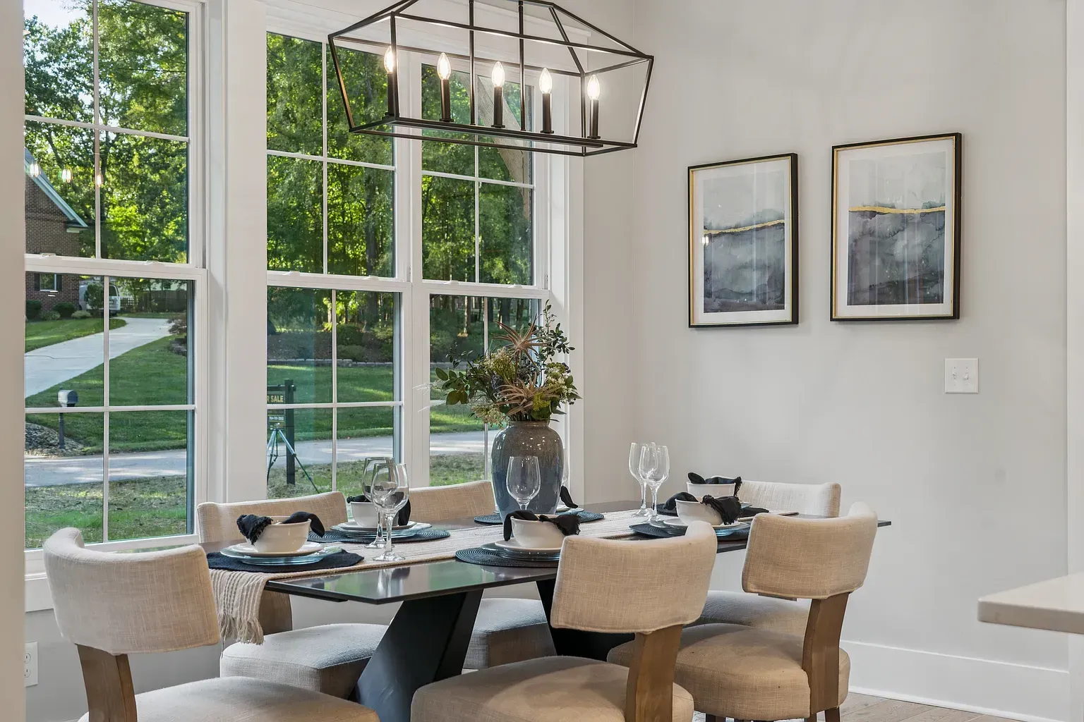 Modern dining room with a glass table, beige chairs, large windows, and framed wall art