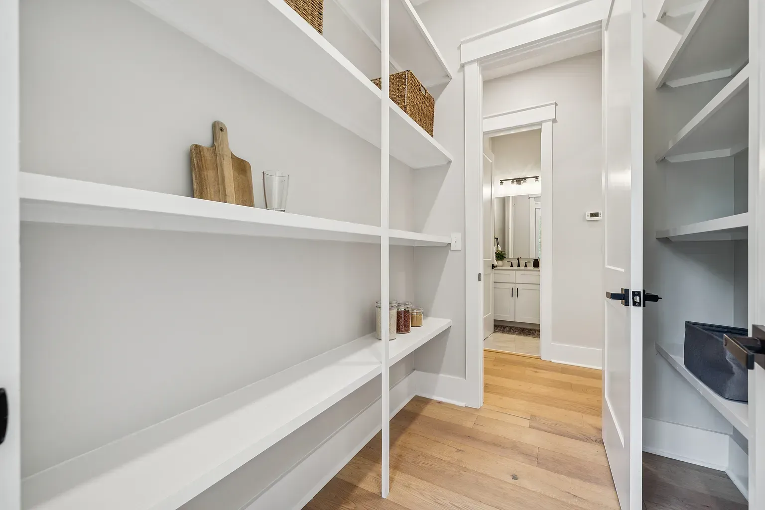 Empty white walk-in closet with built-in shelves and light wood floors, opening to a hallway.
