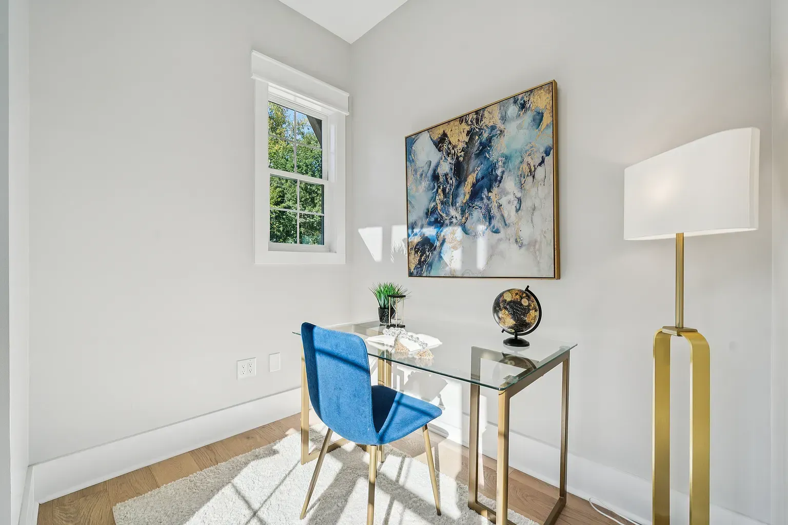 Bright minimalist office with blue chair, glass desk, abstract art, and floor lamp in a sunlit room