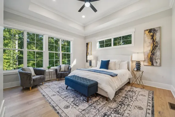 Bright bedroom with large windows, white bed, blue bench, gray chair, and patterned rug.